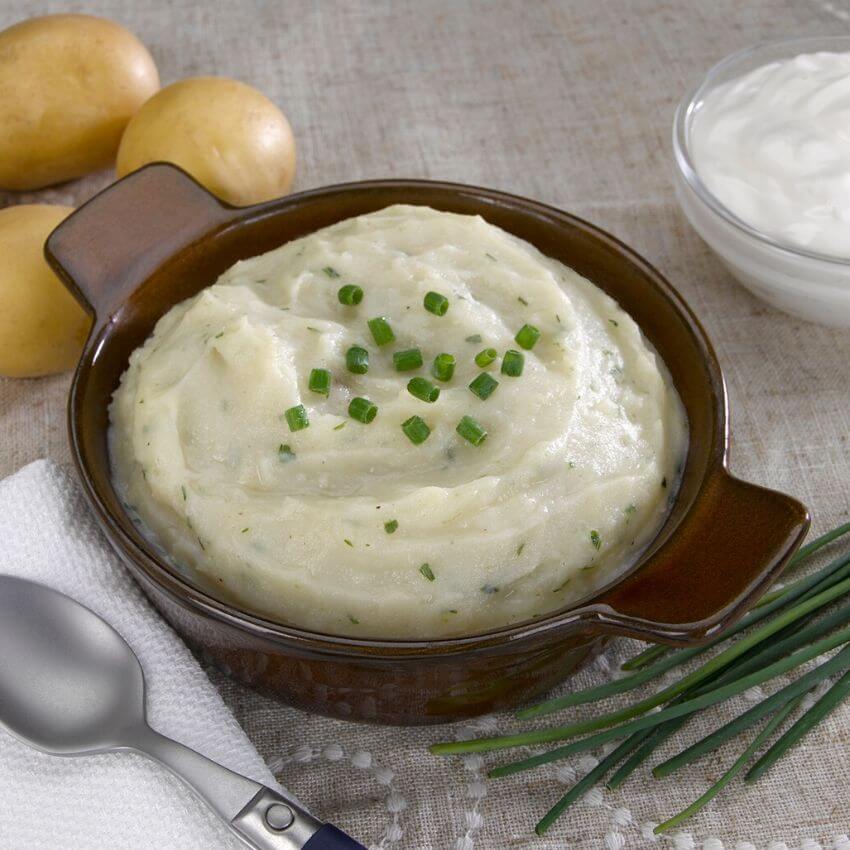 Creamy protein mashed potatoes garnished with chives, served in a brown dish alongside potatoes and sour cream.